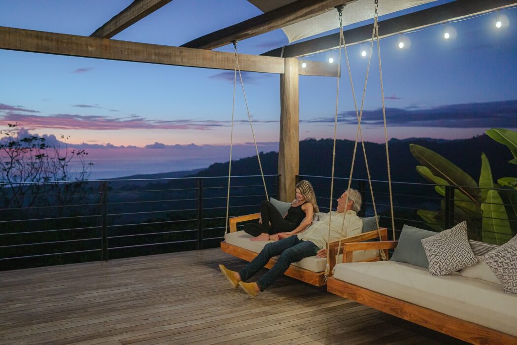 Two people relaxing on suspended wooden daybeds on a modern deck overlooking mountain sunset views with string lights