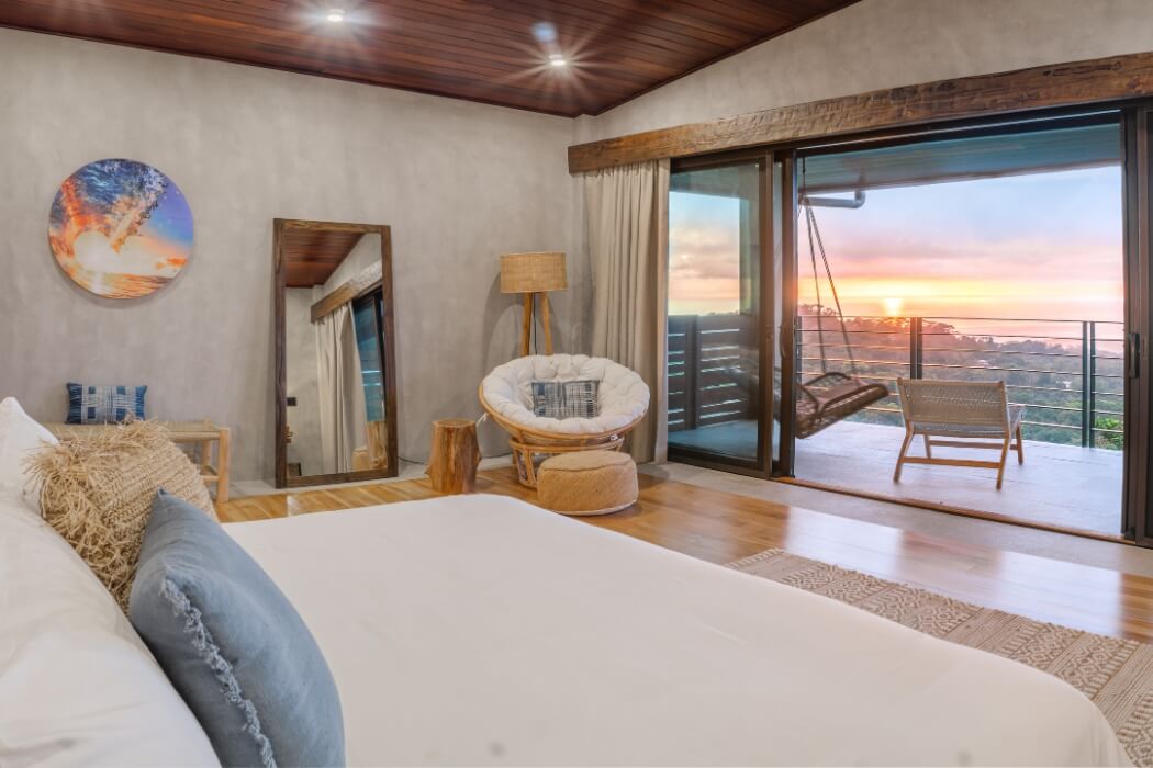 Luxurious bedroom interior with sliding glass doors opening to balcony, papasan chair, and sunset ocean views