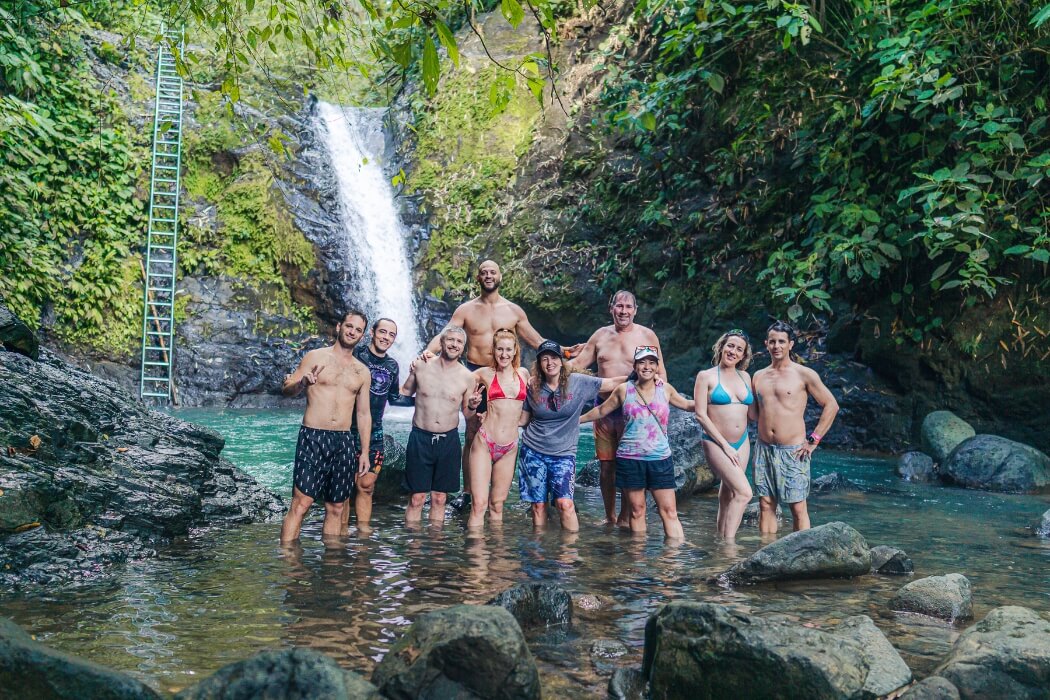 Group of people standing in water at base of tropical waterfall with metal ladder on rock face