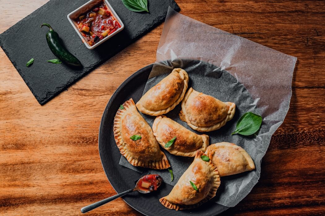 Golden-brown baked empanadas served on black plate with spicy salsa, jalapeño pepper, and fresh basil leaves on wooden surface