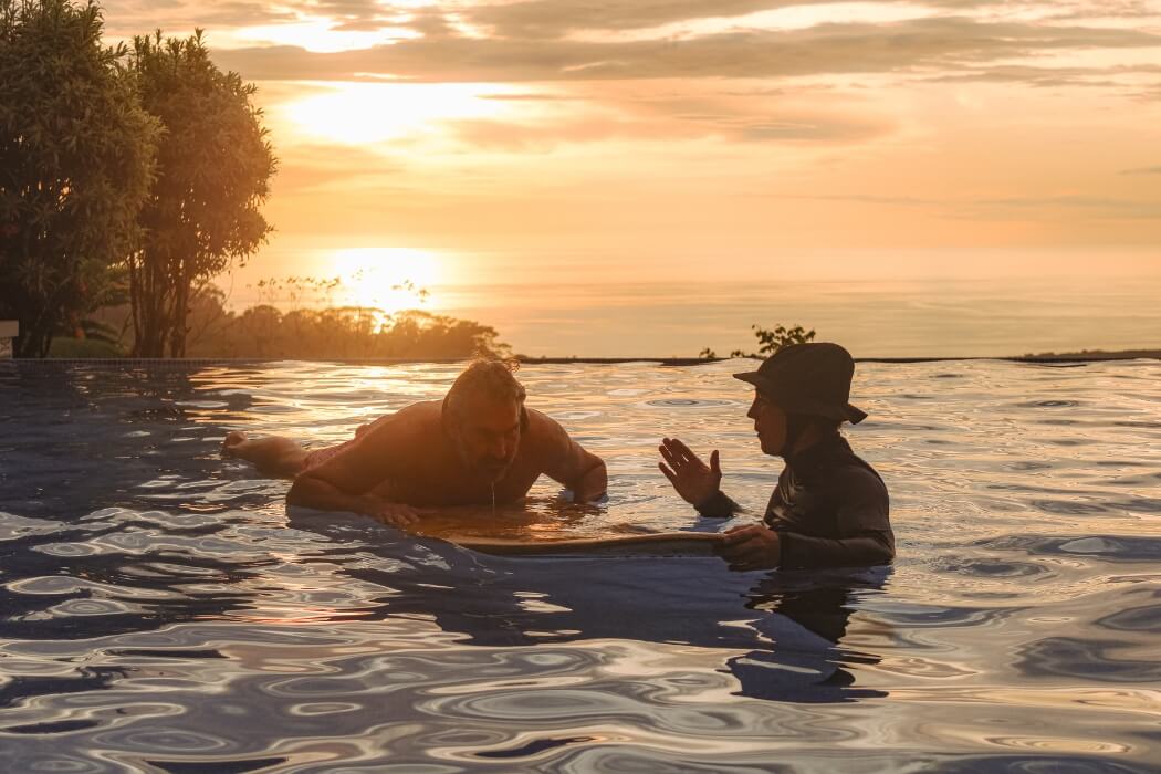 Two people having conversation in infinity pool at sunset with golden sky and ocean view background