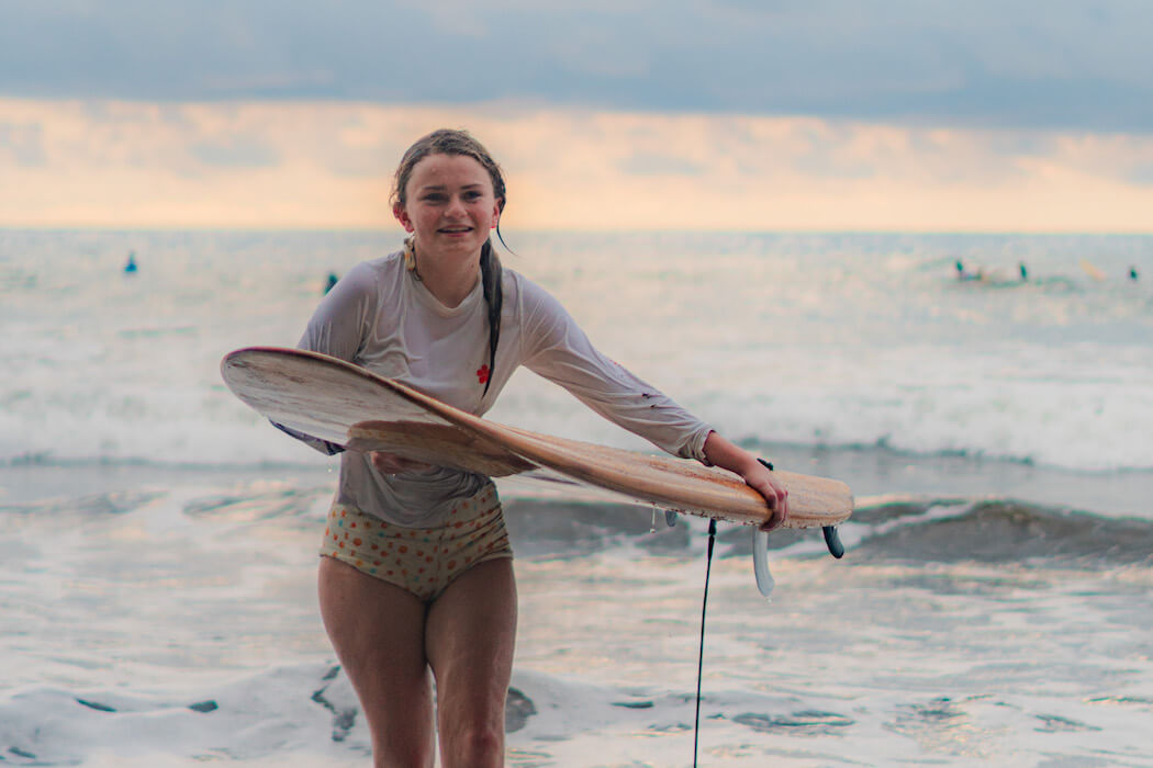 Young woman in rash guard holding surfboard at beach during golden hour with waves in background