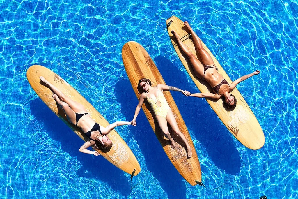 Surf Sisters Under the Sun Top view of three women lying on their surfboards, floating side by side in a clear blue pool, holding hands and soaking up the sun, creating a peaceful and joyful summer vibe.