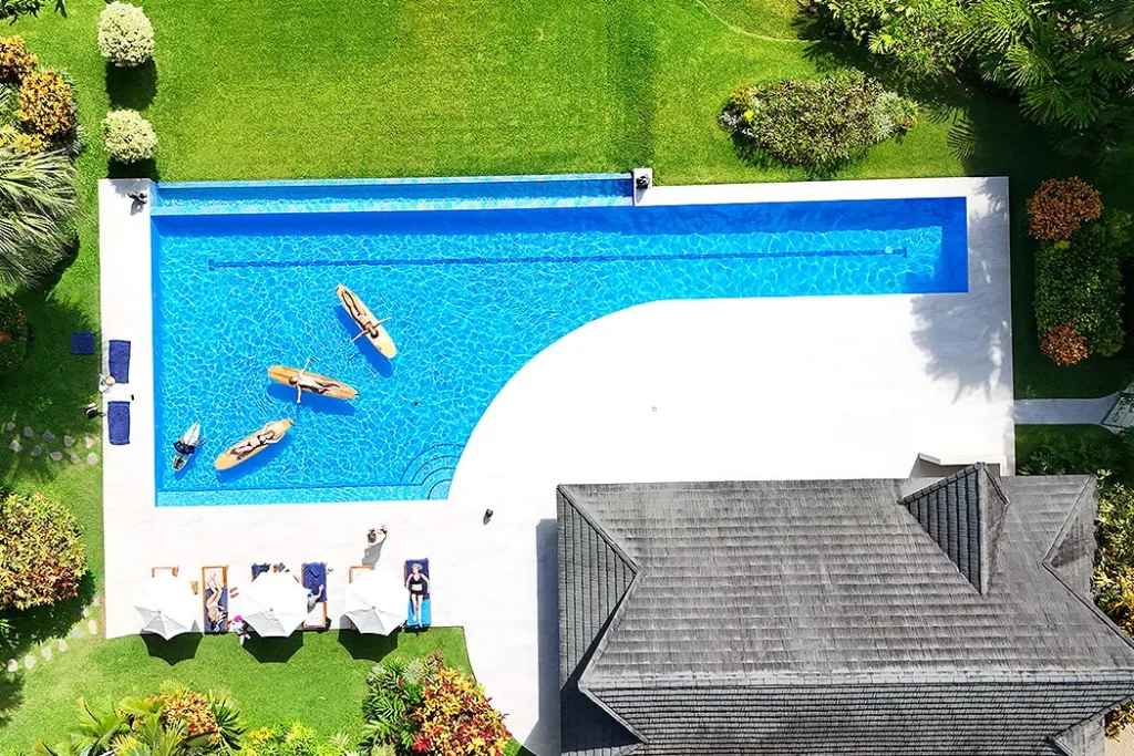 Sun-Soaked Serenity at the Resort Zoomed-out top view of three women lying on surfboards, floating in a large resort pool while holding hands. The surrounding scene includes the resort’s roof, green grass fields, and trees, capturing a tranquil and scenic summer moment.