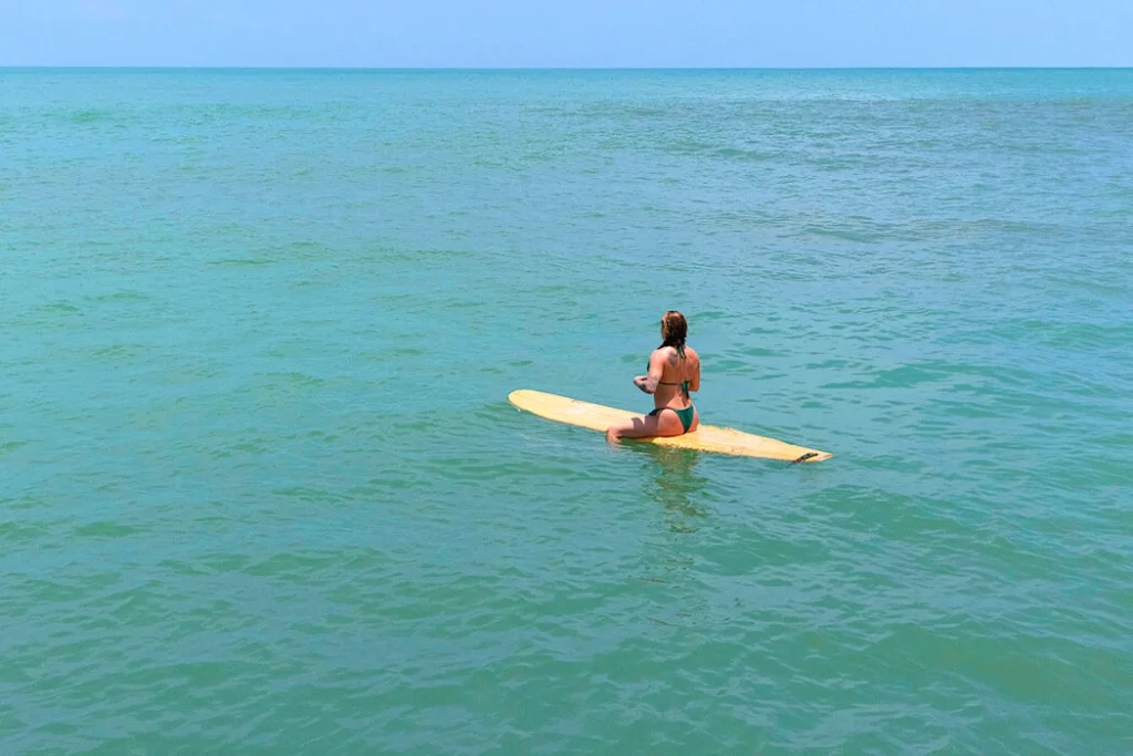 Surfer Woman Amidst the Vast Sea A woman sitting on her surfboard floating in the middle of the vast sea, captured in an environmental shot that highlights the expansive water surrounding her, emphasizing the scale between the surfer and the ocean.
