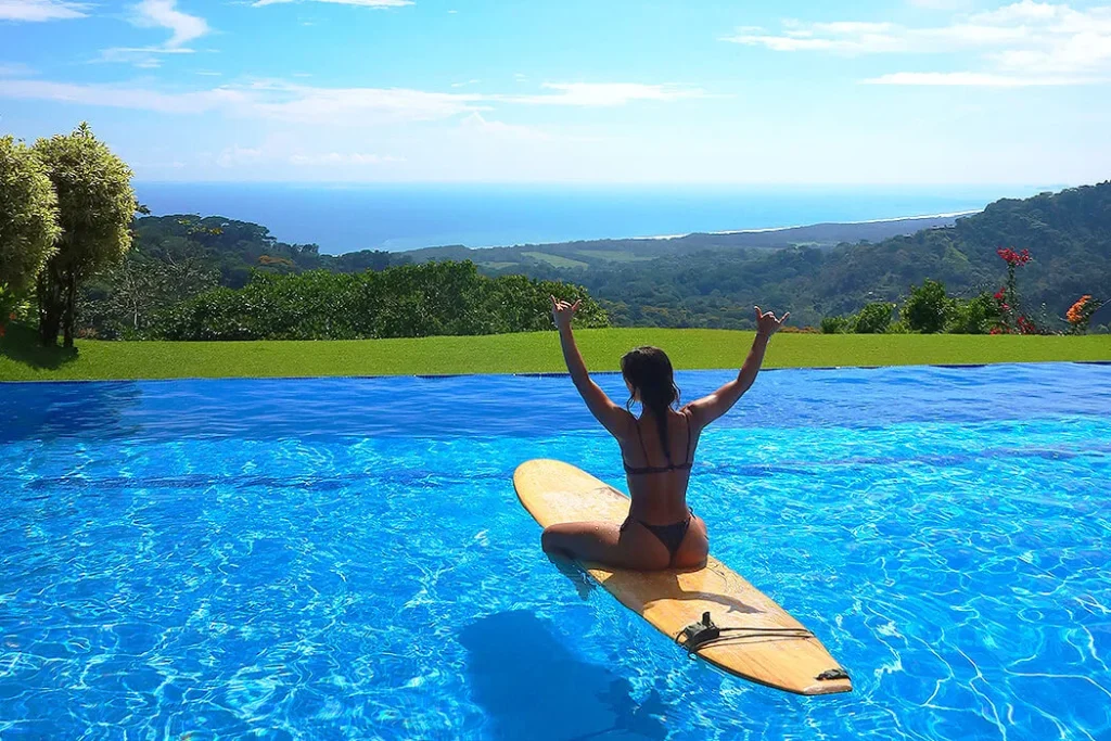 A woman sitting on her surfboard floating in a resort pool, enjoying the scenic view of a lush valley and surrounding trees, creating a peaceful and serene atmosphere.