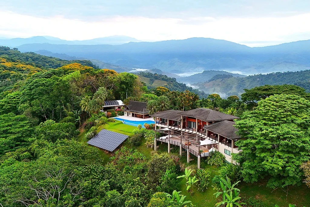 Mountain Villa Resort with Solar-Powered Elegance Environmental aerial shot of a beautiful villa resort perched atop a mountain hill, surrounded by hills, mountains, and dense trees. The resort’s pool is visible, and solar panels on the roofs highlight its sustainable energy use.