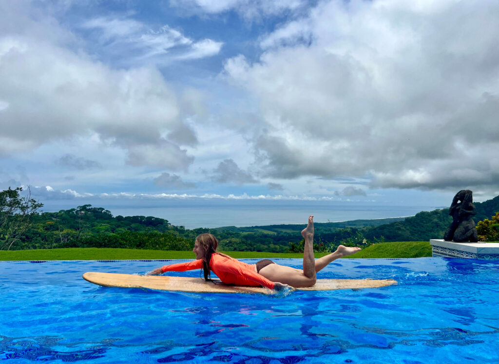 Surf Training in Pool at Lamangata Resort – Woman Practicing on Floating Board Woman in a tropical resort pool at Lamangata, practicing pop-up technique on a floating surfboard, surrounded by lush greenery.
