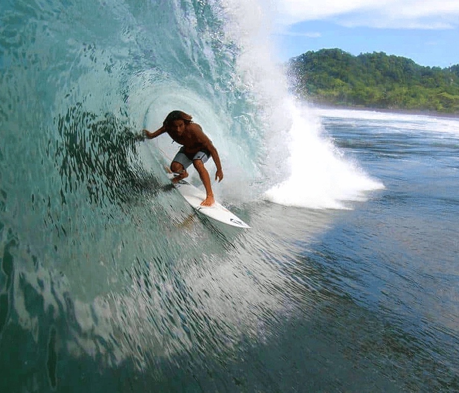 Experienced surfer riding a long, peeling left-hand wave at Pavones, Costa Rica, with jungle-lined coastline in the background.