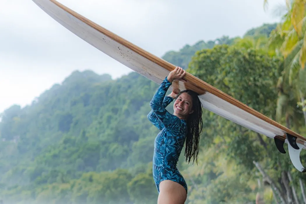 Woman in a blue rash guard holding a long surfboard overhead on a tropical beach, with lush green hills and palm trees in the background