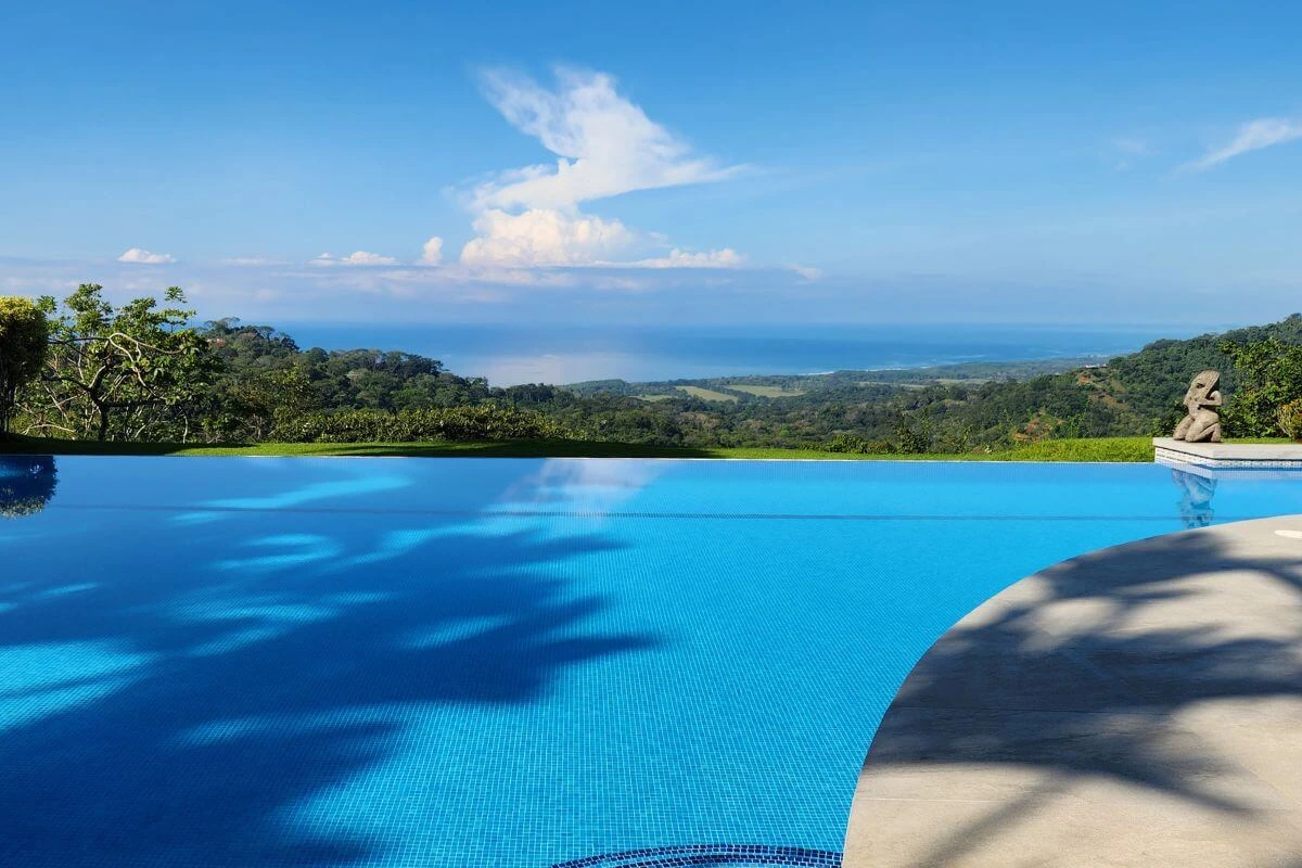 Bright midday view of a serene infinity pool reflecting the sky, with lush green hills and the ocean on the horizon, palm shadows on the deck, and a stone statue at the corner.