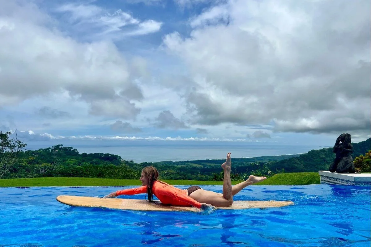 Woman in an orange rash guard lies on a surfboard in a bright blue infinity pool, practicing paddling with one leg raised, overlooking lush green hills and the ocean under a cloudy sky, with a dark statue on the pool edge to the right.