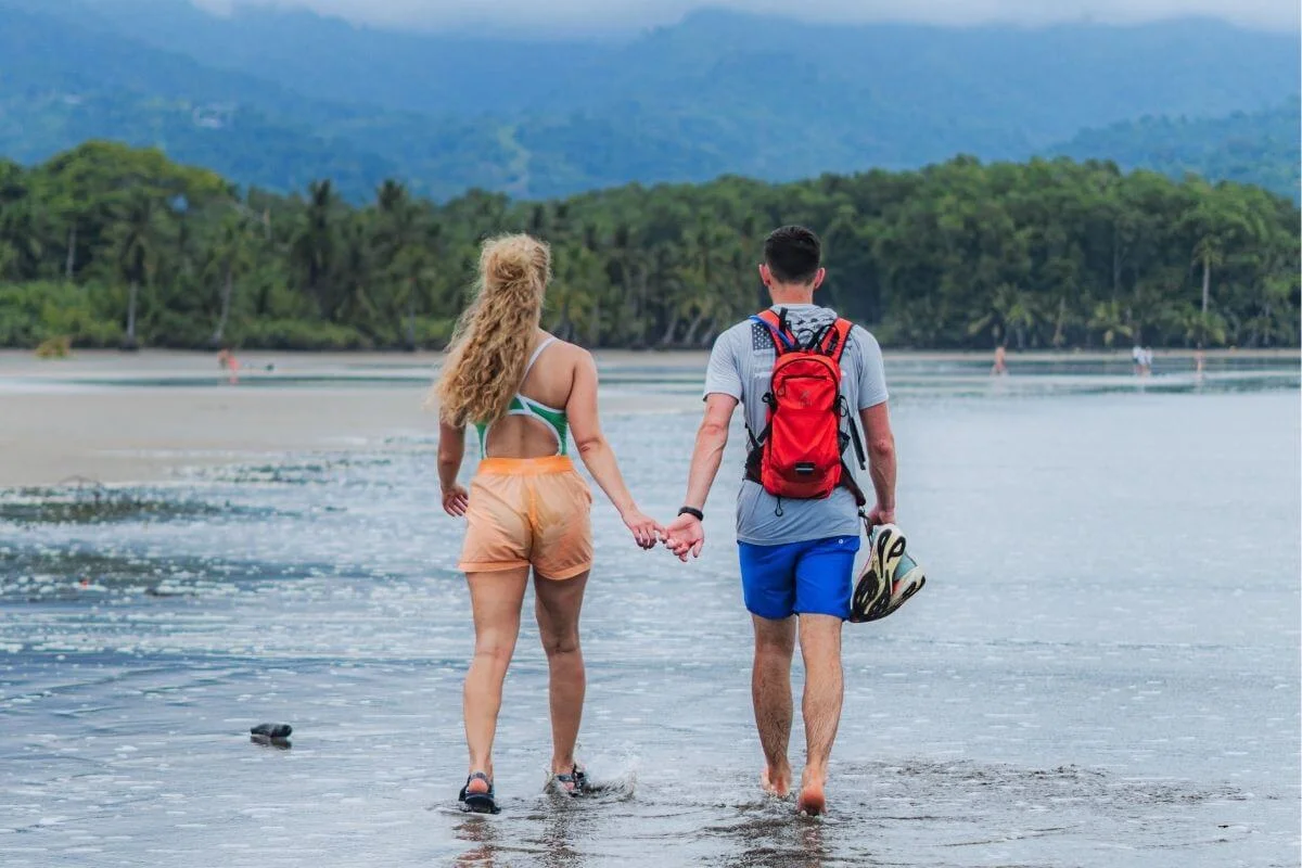 Rear view of a couple holding hands while walking along a shallow, reflective shoreline toward a tropical tree line and misty mountains, with the man carrying sandals and wearing a red backpack.