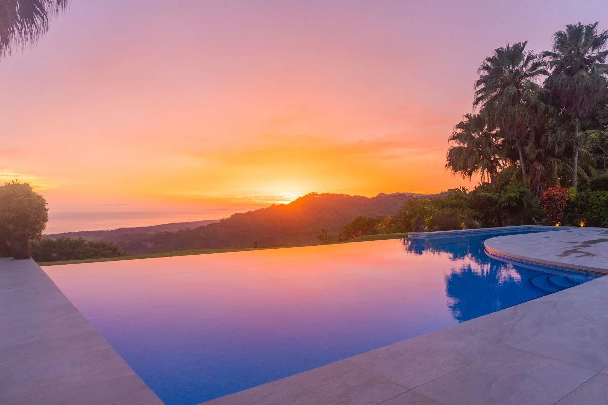 Tranquil infinity pool reflecting pink and orange sunset skies, overlooking lush green hills and distant ocean, framed by palm trees and landscaped gardens on the right.