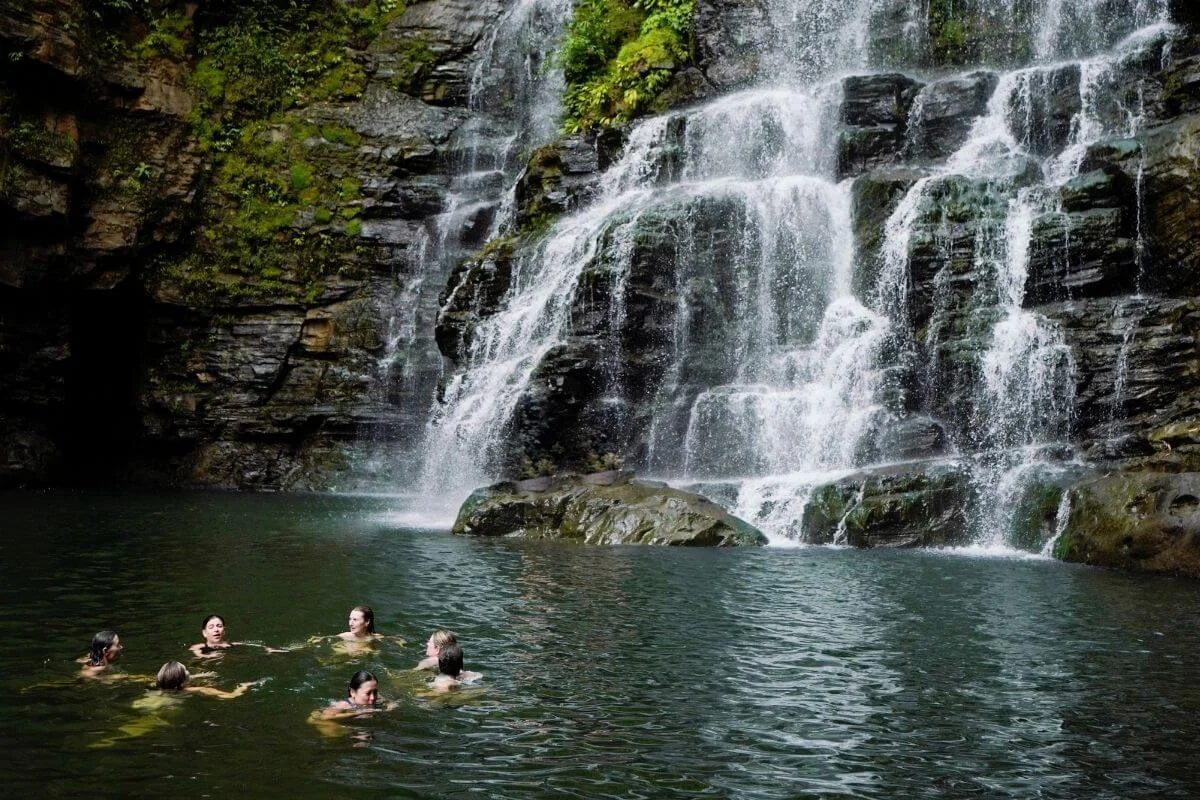 Several people swim together in a calm green pool at the base of a tall, rocky waterfall, surrounded by moss-covered cliffs and lush vegetation.
