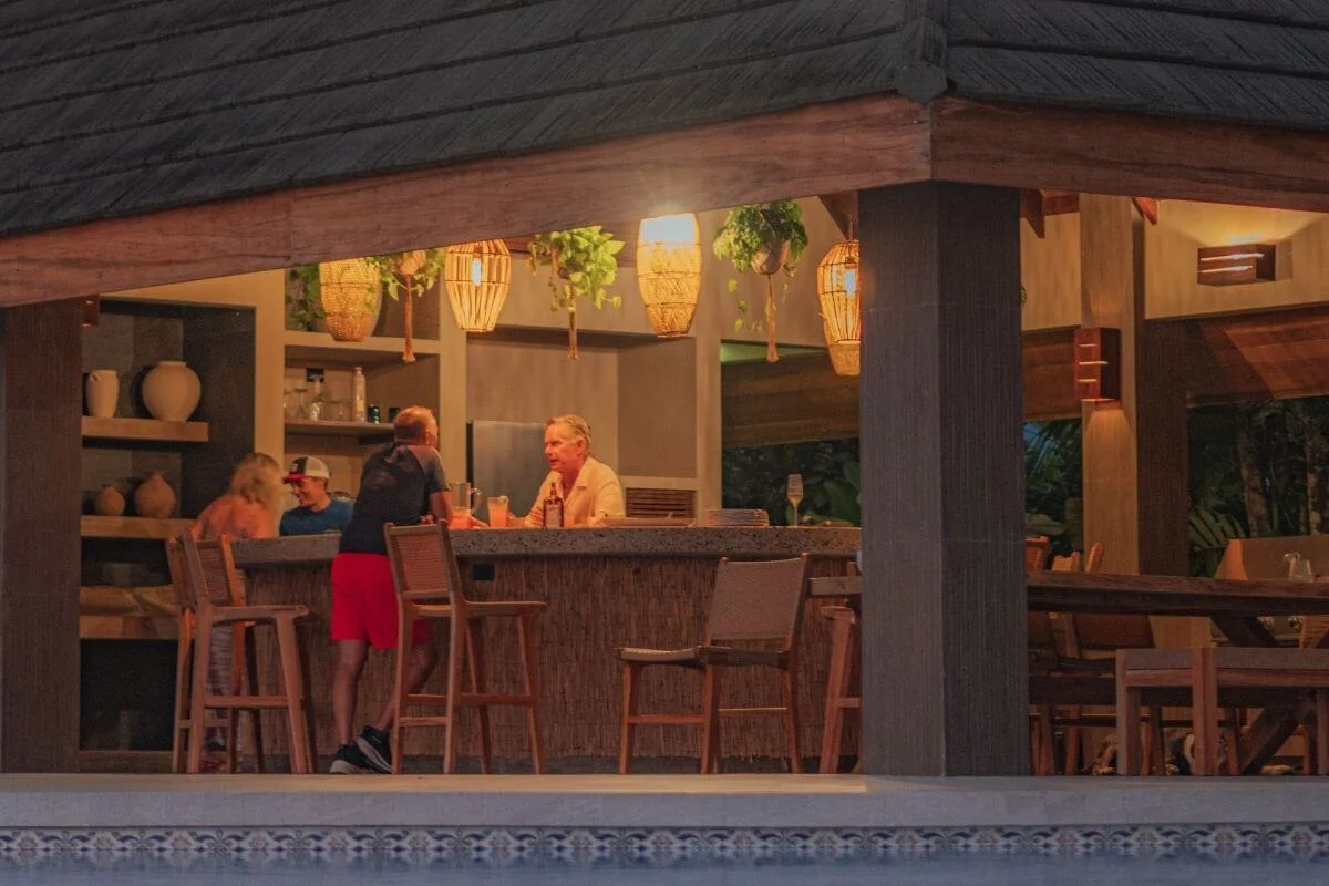 A small group of guests sit and chat at a warmly lit open-air bar with wooden stools, hanging plants, and woven lanterns, next to a pool in a tropical resort.