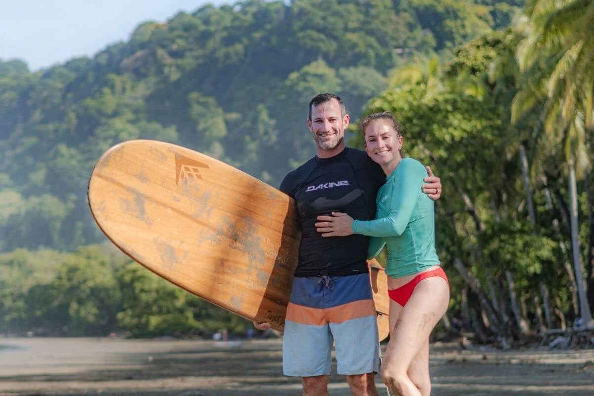 A smiling couple in swimwear and rash guards stands on a sandy tropical beach, hugging beside a large wooden surfboard, with lush green hills and palm trees in the background.