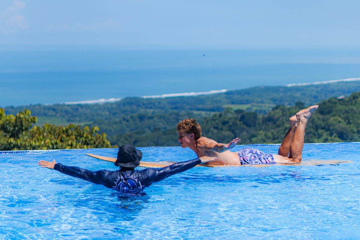 Instructor and surfer practicing balance on a surfboard in an infinity pool with ocean and tropical landscape views in the background