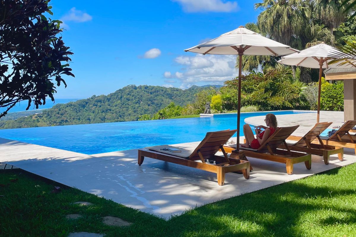Woman relaxing on a lounge chair beside an infinity pool with lush green mountains and ocean views in the background
