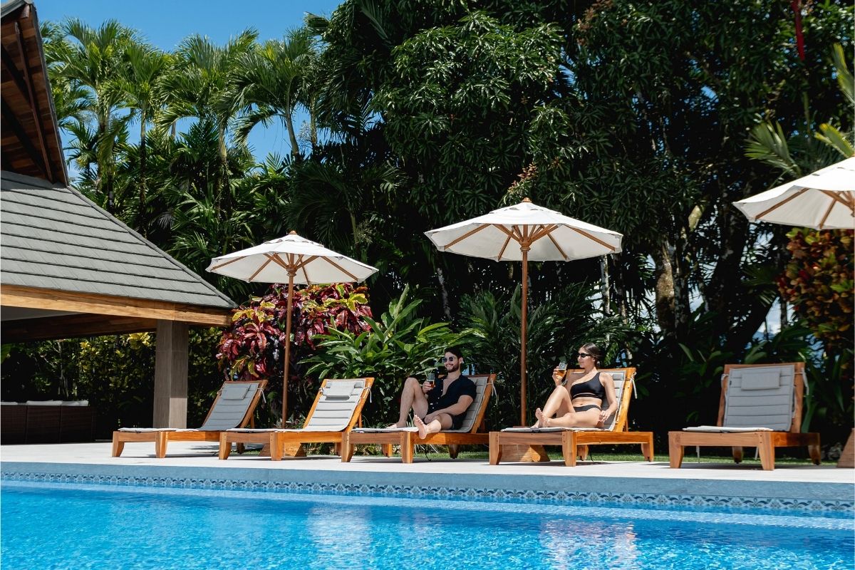 Two guests lounging on sunbeds by a tropical pool with umbrellas, surrounded by lush greenery at a luxury resort in Costa Rica.
