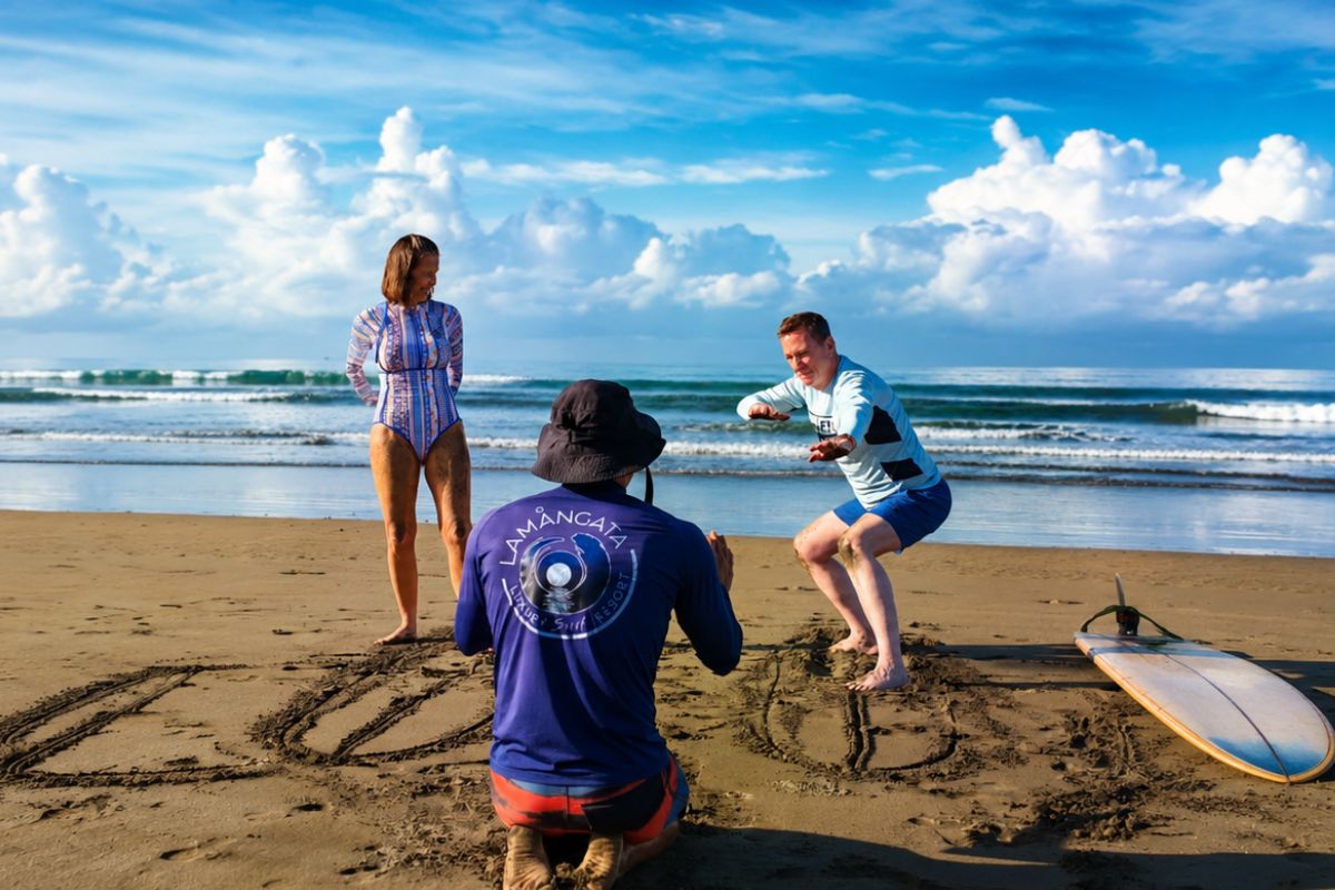 A Lamangata surf coach in a branded blue shirt kneels on the sand to demonstrate surfing technique to two guests, with a surfboard beside them, waves breaking in the background, and dramatic clouds over the tropical coastline.