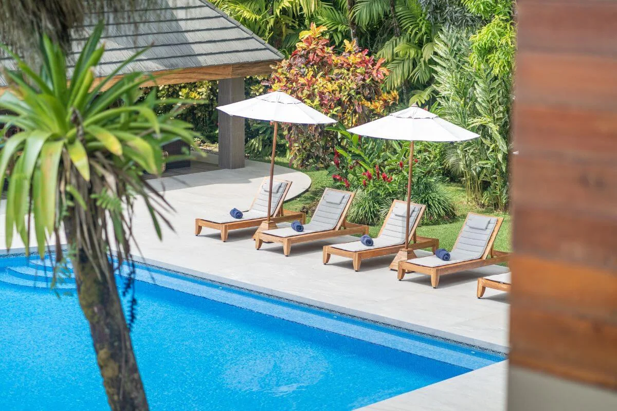 Four teak lounge chairs with white umbrellas beside a blue pool surrounded by tropical gardens