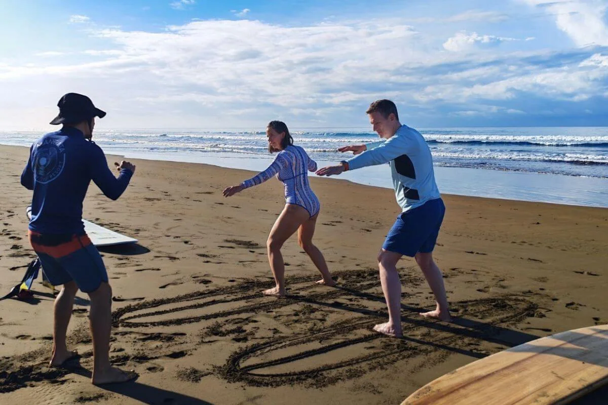 A Lamangata surf instructor in a branded blue shirt coaches two guests practicing their surf stance on the sand, with surfboard outlines drawn in the sand beneath them, breaking waves and a dramatic cloudy sky in the background.