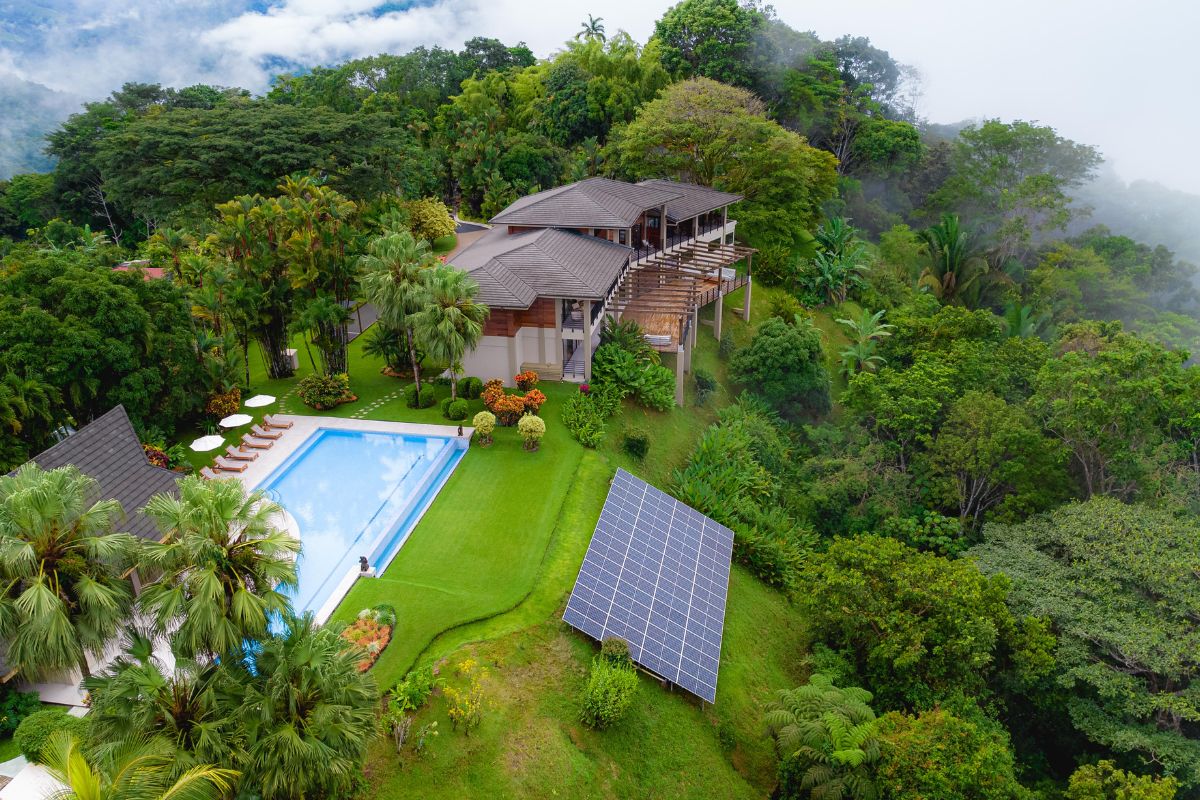 Drone view of the Lamangata surf lodge nestled in lush tropical rainforest, featuring a large rectangular pool with sun loungers and umbrellas, a multi-level wood and concrete main building with a wraparound deck, and a solar panel array on the grounds below.