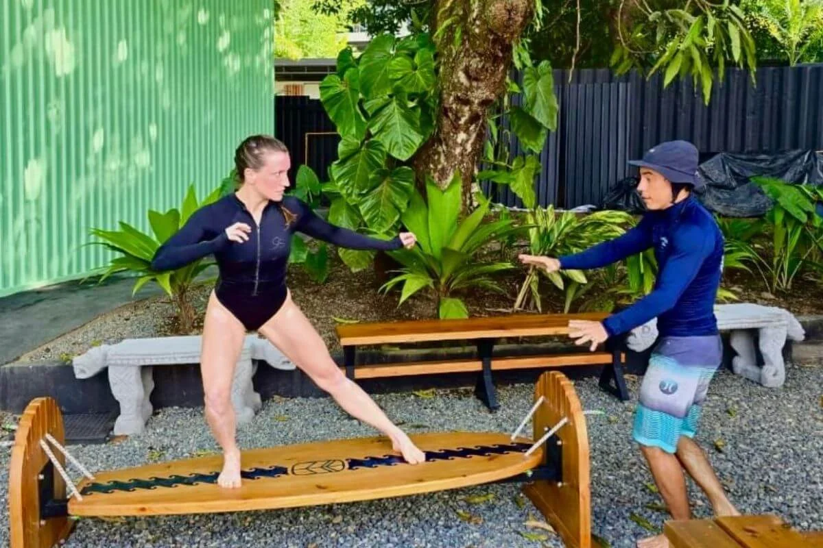 Surf instructor guiding a woman practicing stance on a wooden balance board outdoors