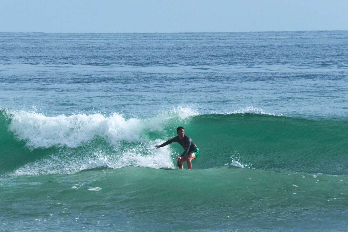 A surfer in a gray rash guard and teal shorts carves across a rolling green wave with white water breaking behind, set against a calm blue ocean under clear daylight.