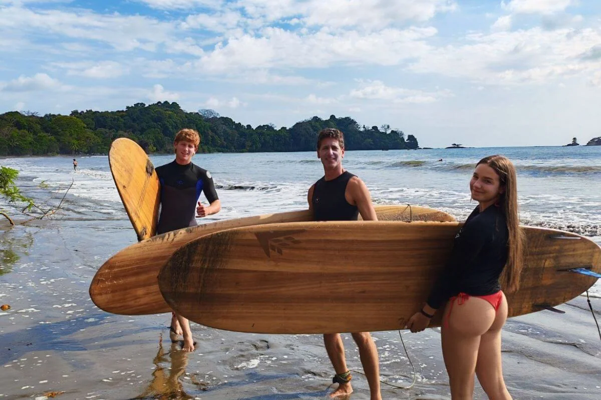 Two young men and a woman stand smiling on a tropical beach holding large natural wooden surfboards, with gentle waves breaking behind them and a densely forested headland visible across the bay under a partly cloudy sky.