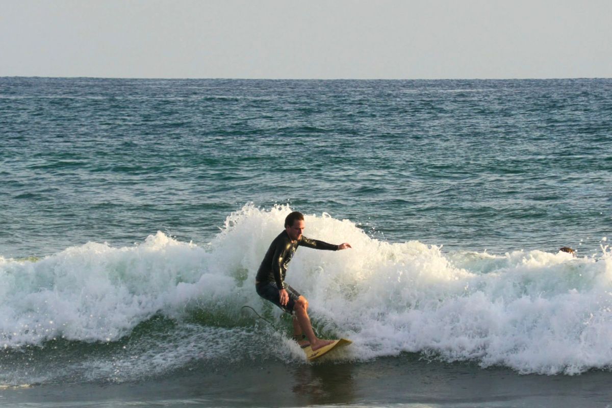 A surfer in a black wetsuit rides a crashing white wave low and balanced on a yellow board, with another surfer visible in the water behind and a calm blue-green ocean in the background.