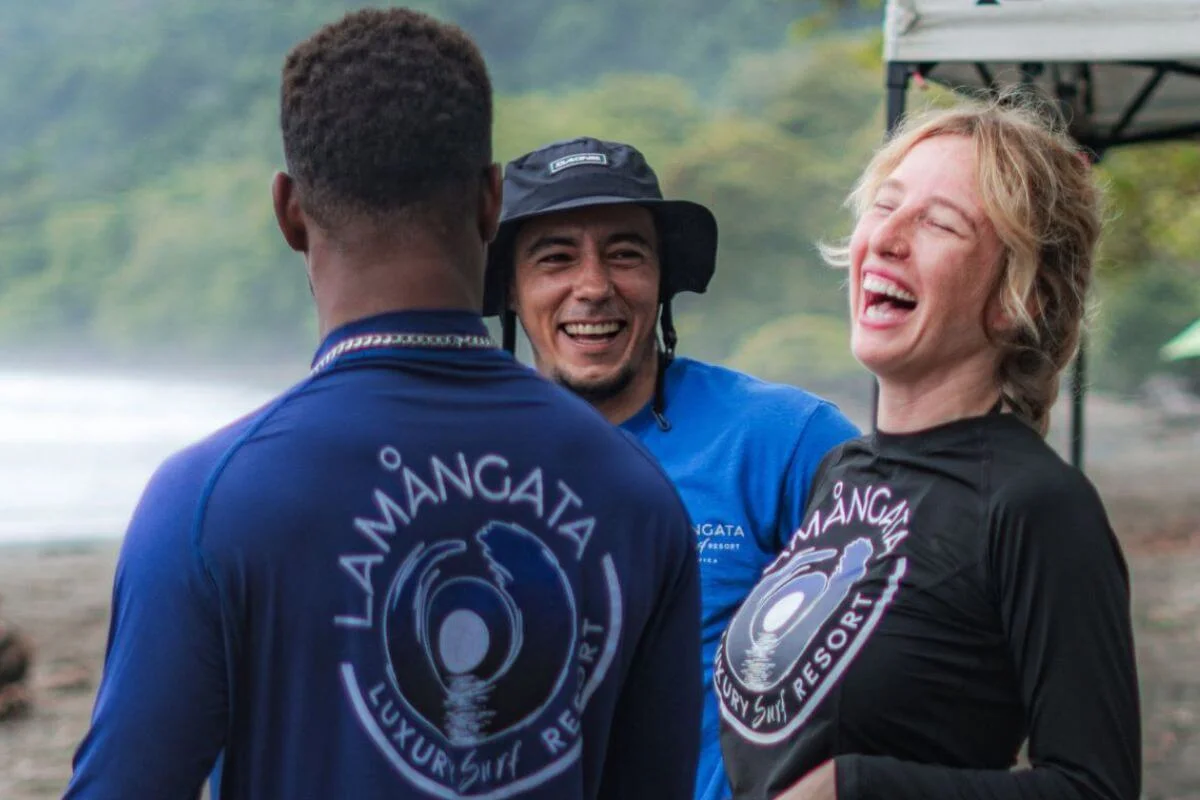 Two Lamangata surf instructors in branded rash guards share a laugh with a female guest on the beach, with a misty jungle coastline visible in the background, capturing the warm and welcoming atmosphere of the resort.