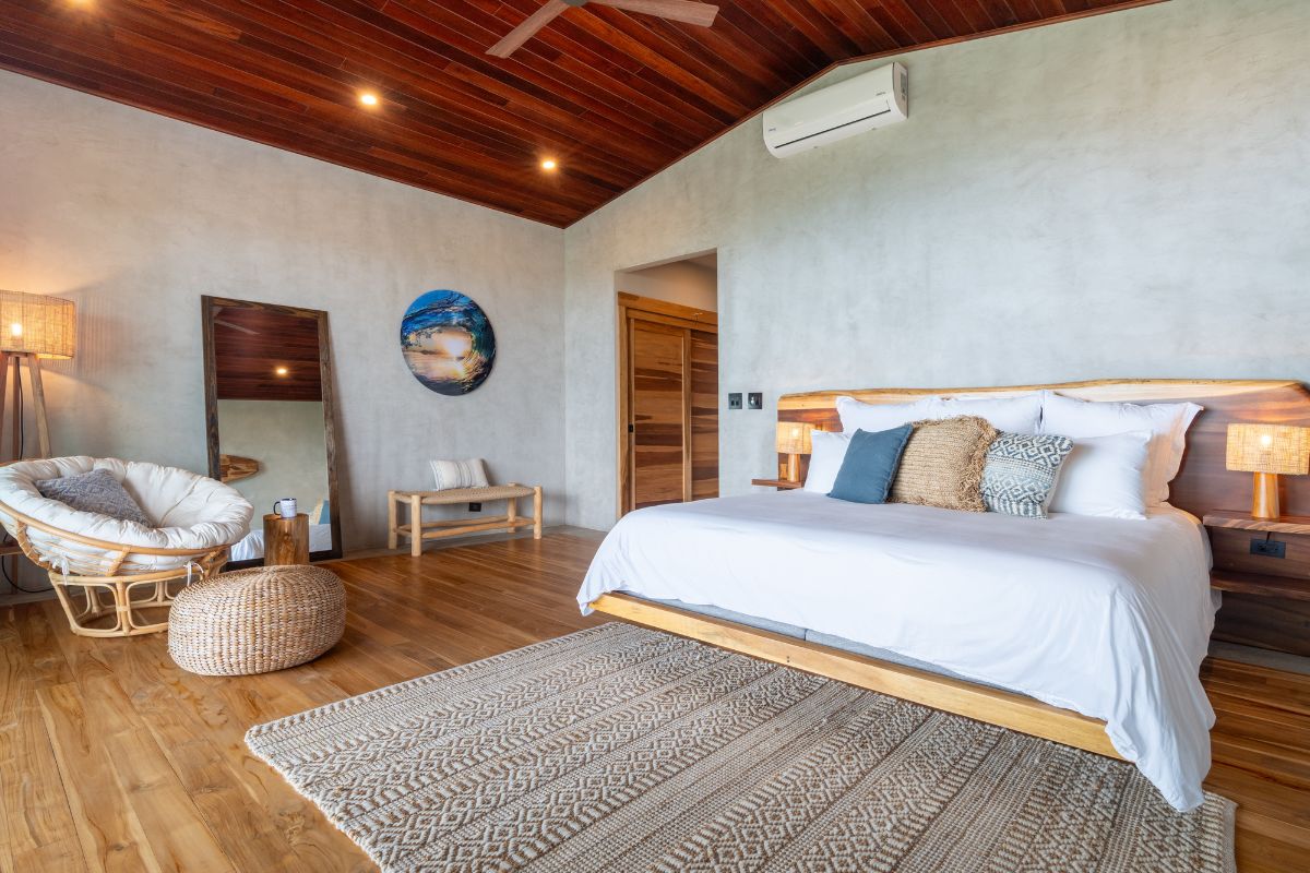 A bright, airy hotel suite featuring a king bed with white linens and layered textured cushions on a raw wood frame, a rattan papasan chair, woven ottoman, full-length mirror, and a circular ocean wave artwork on the concrete wall, with warm wood floors and soft lamp lighting throughout.