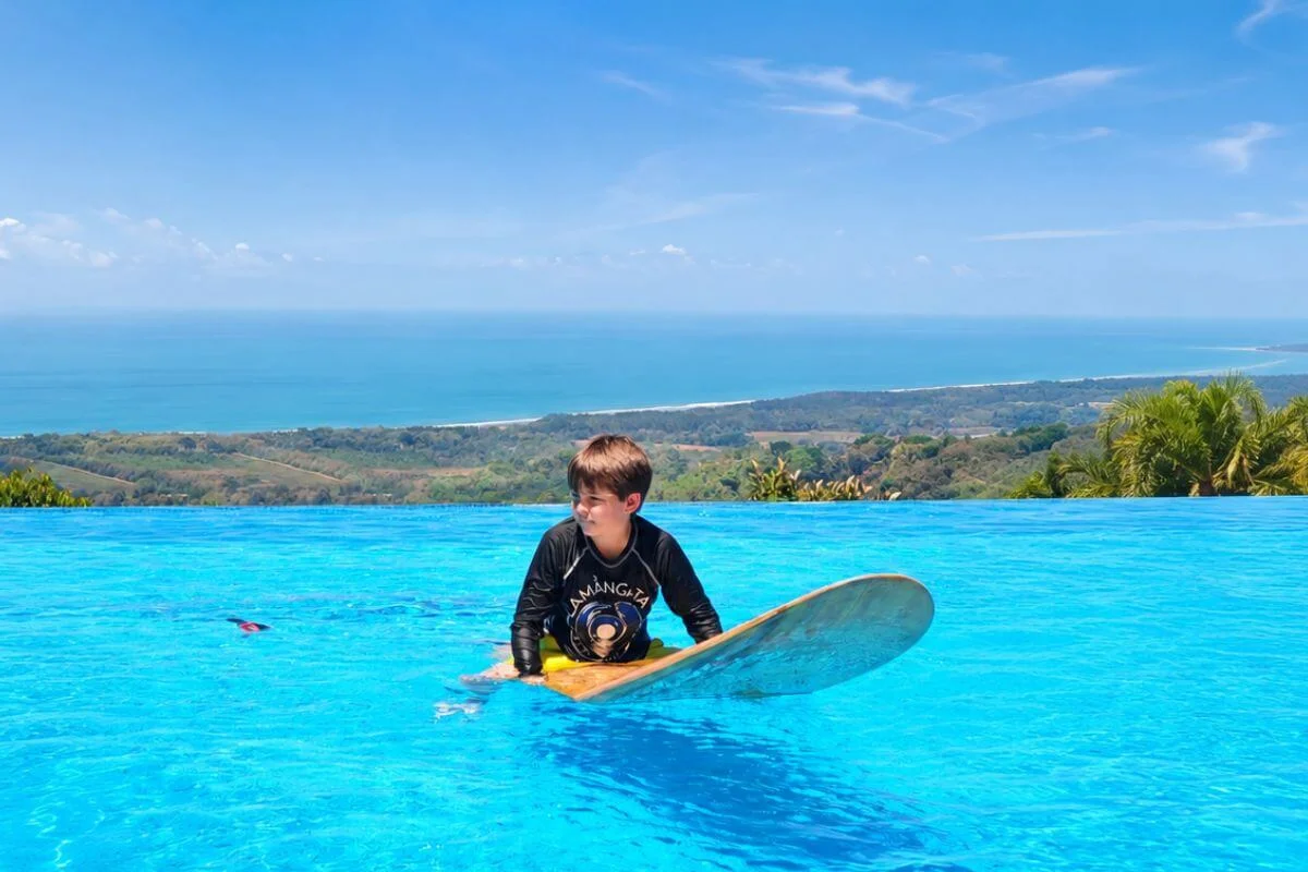A young boy in a Lamangata rash guard lies on a wooden surfboard in the bright blue infinity pool, with a sweeping panoramic view of the Costa Rica coastline, rolling green hills, and the Pacific Ocean stretching to the horizon under a clear blue sky.