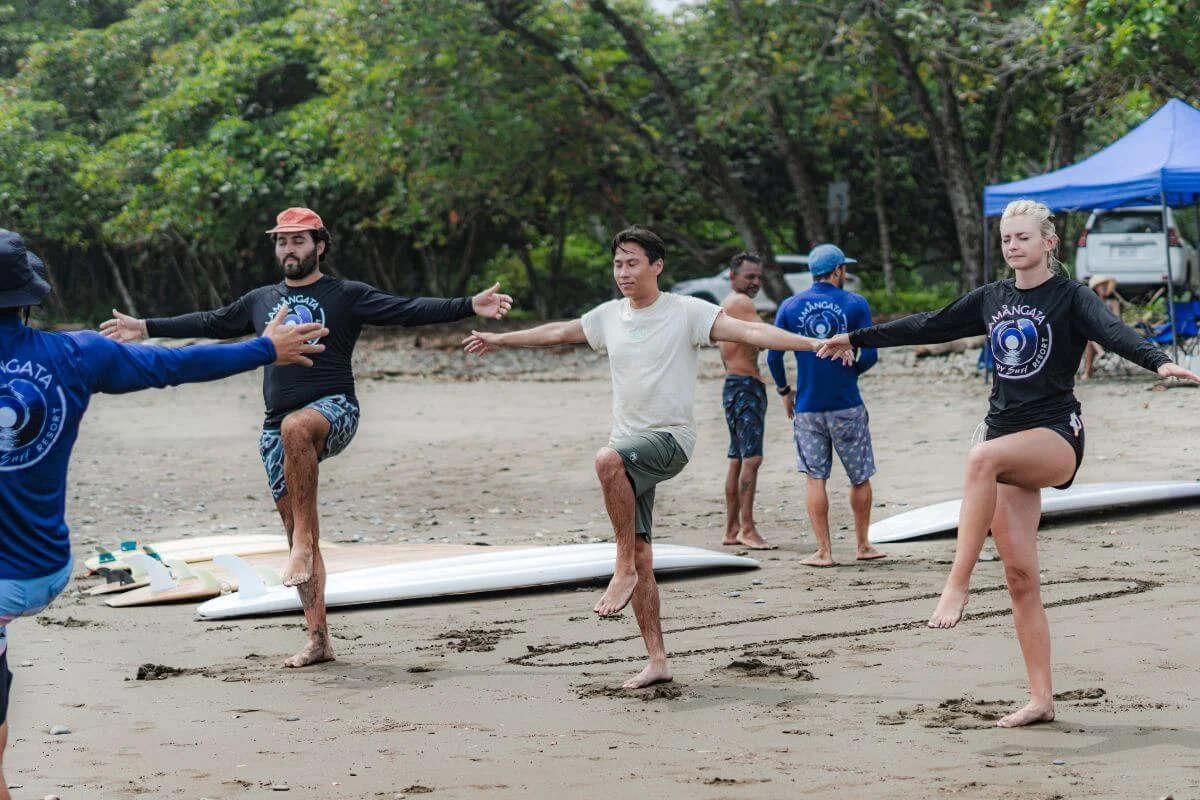 A group of surf guests and Lamangata instructors practice single-leg balance drills with arms outstretched on a sandy beach, with longboards laid out on the sand behind them, a blue canopy tent, and dense tropical trees in the background.