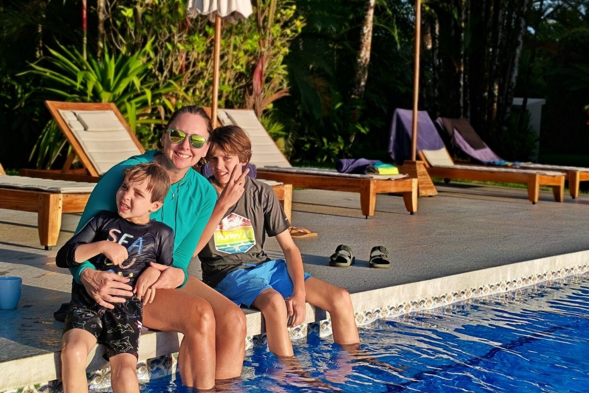 A smiling mother in a teal rash guard sits at the edge of the Lamangata pool with her two young sons, with wooden sun loungers and lush tropical gardens visible in the background at golden hour.