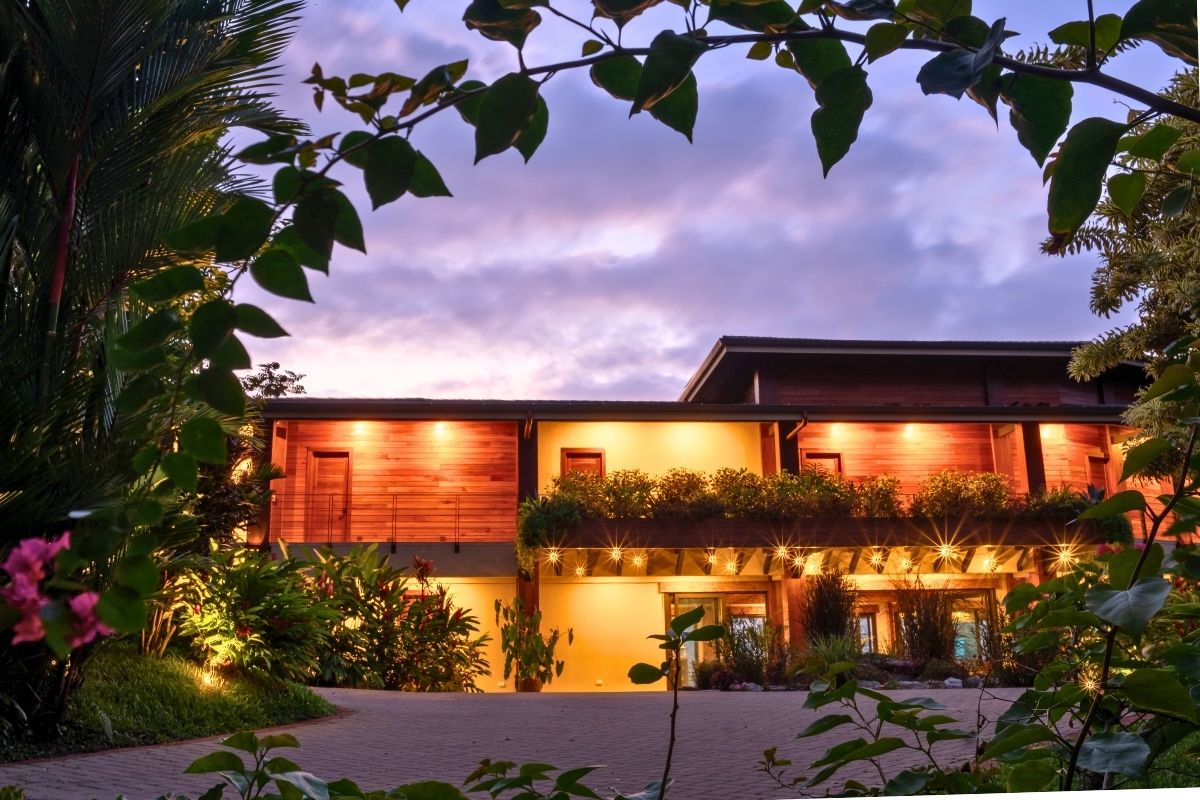 The warm wood and concrete exterior of the Lamangata surf resort glows with golden accent lighting at dusk, framed by tropical foliage and bougainvillea flowers under a dramatic purple and blue sky.