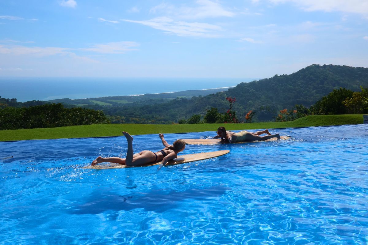 Two women lie on wooden surfboards in the Lamangata infinity pool, with lush green hills, a white sand coastline, and the Pacific Ocean stretching to the horizon under a bright blue sky.