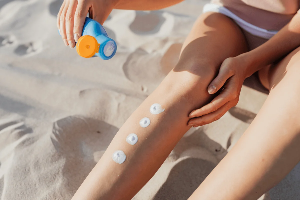 Person applying sunscreen from a blue bottle onto their leg while sitting on a sandy beach