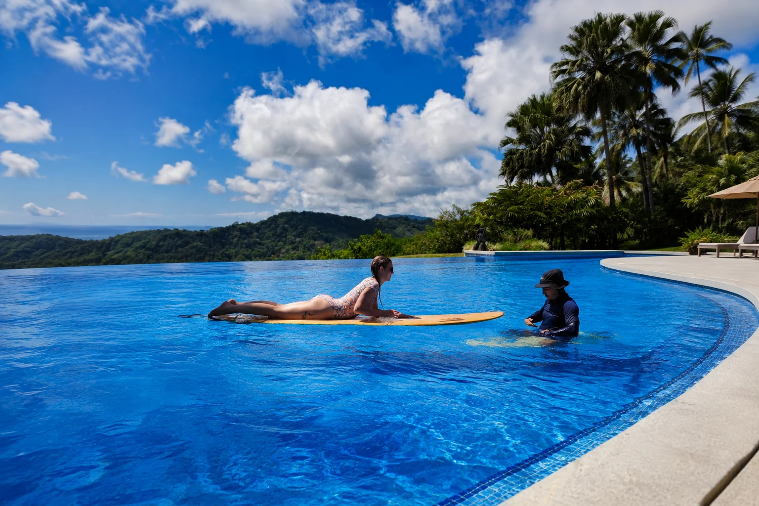Surf instructor teaching a woman to paddleboard on a longboard in a tropical resort pool, with lush mountains, palm trees, and a bright blue sky in the background.