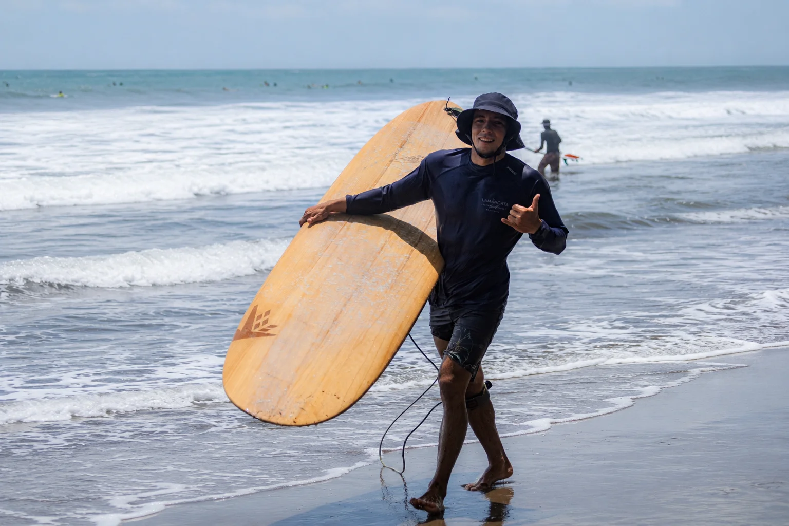 Surf instructor at Lamangata carrying a wooden longboard on the beach, giving a shaka sign