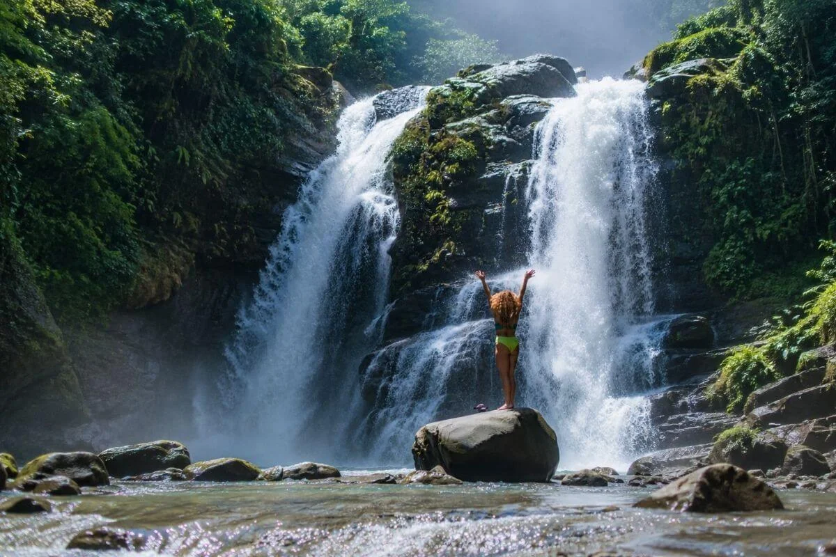 A woman standing on a large rock in a bikini with her arms raised, looking up at a powerful cascading waterfall surrounded by lush tropical rainforest.