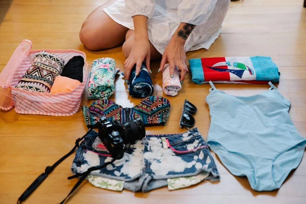 Beach vacation essentials arranged on a wooden floor, including rolled clothing, swimwear, sunglasses, a camera, and a travel bag.