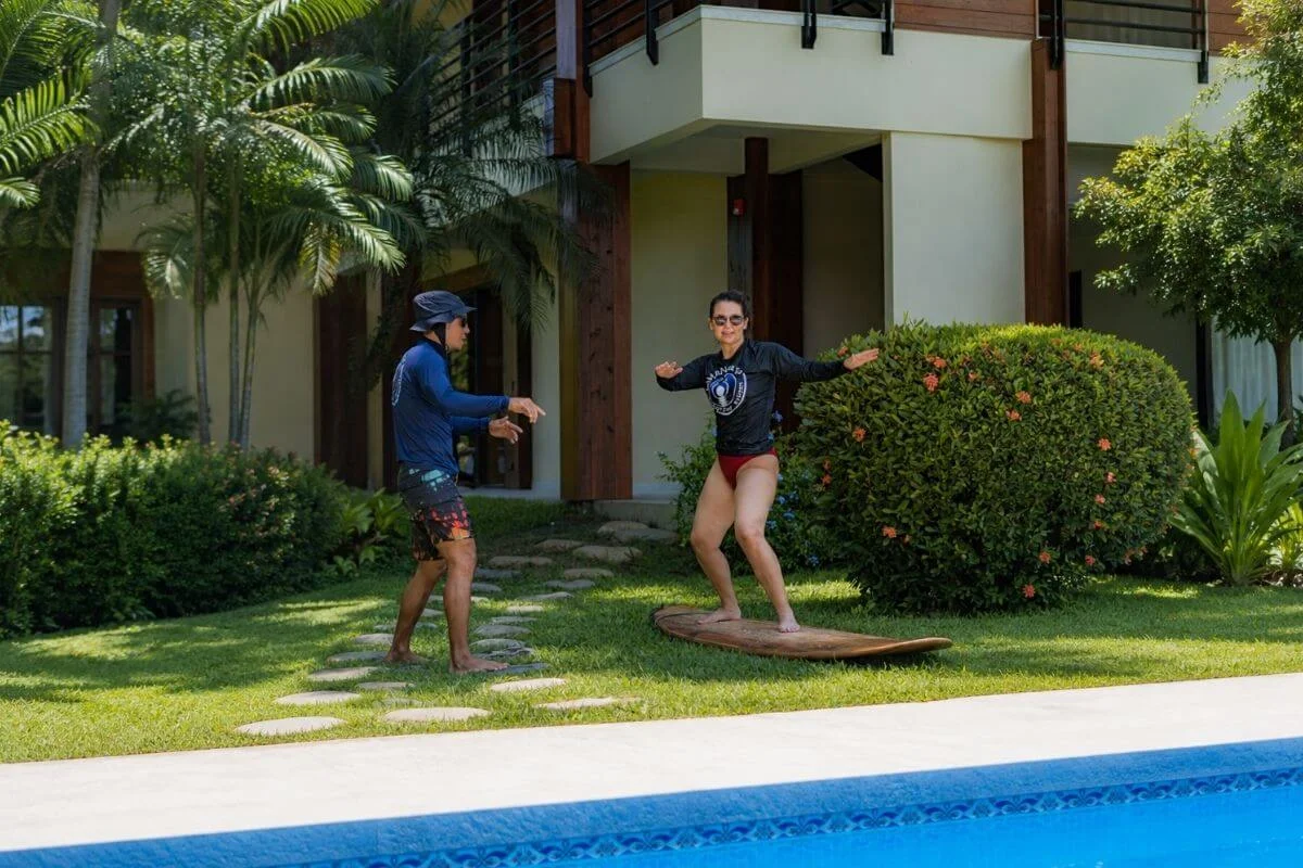 An instructor teaching a woman how to balance on a surfboard on the grass near a swimming pool and tropical resort villa.