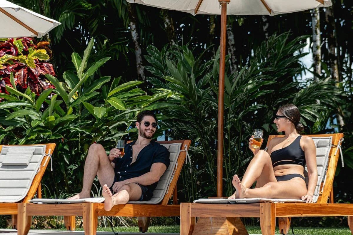 A man and woman relaxing on wooden sun loungers under an umbrella, holding drinks surrounded by dense green tropical plants.