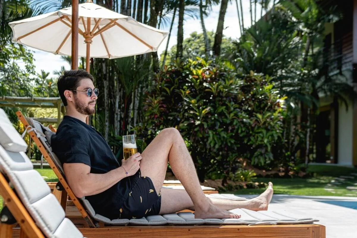 A man relaxing on a wooden sun lounger under a white umbrella, holding a drink poolside with tropical foliage in the background.
