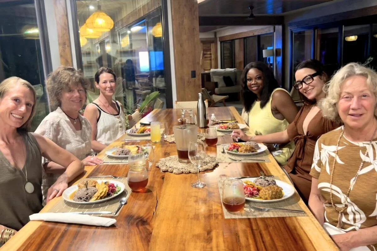 A group of women sitting together at a long wooden dining table enjoying traditional Costa Rican meals and drinks inside a brightly lit lodge.