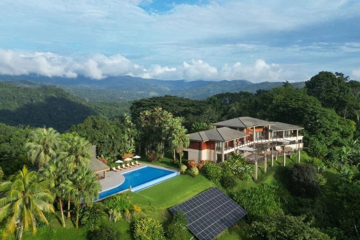 Drone shot of a luxury eco-lodge with a swimming pool, solar panels, and outdoor deck nestled in a dense tropical rainforest facing mountain hills.