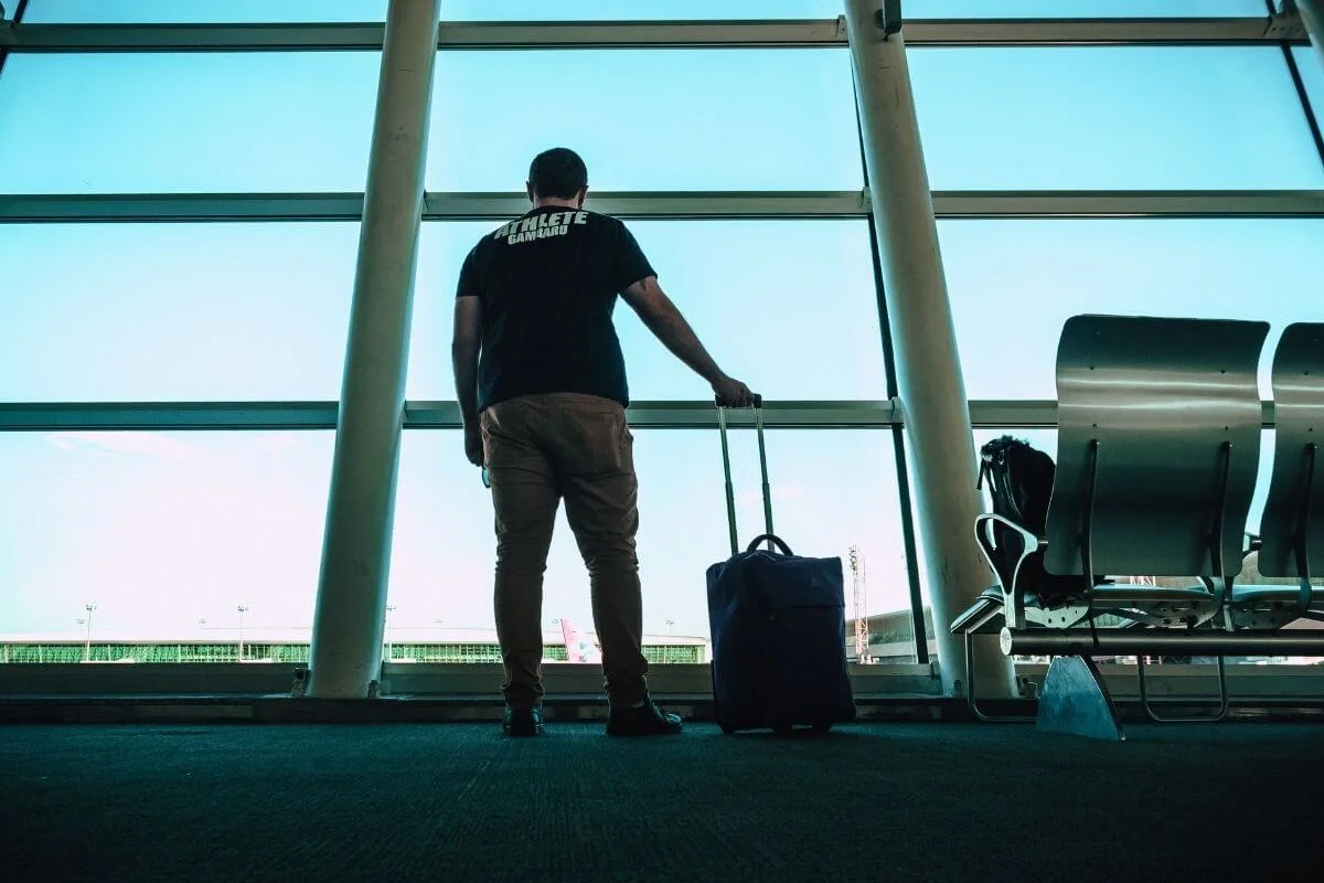 A male traveler with a rolling suitcase standing in front of large glass windows inside an airport departure terminal looking out at the tarmac.